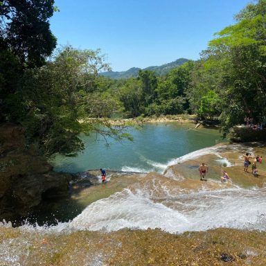 Roberto Barrios Cascade d'eau claire avec des personnes se baignant dans une piscine naturelle entourée de verdure.